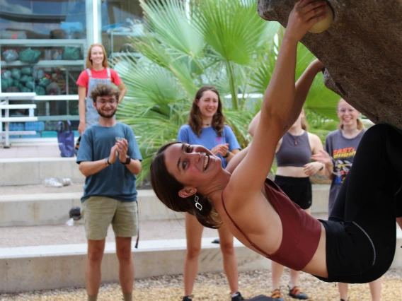 A climber hangs from a bouldering wall with people smiling in background