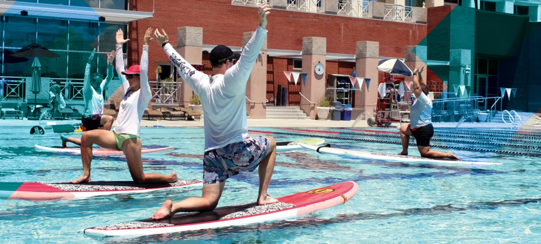 Several young people doing yoga poses on top of floating paddleboards in a university pool.