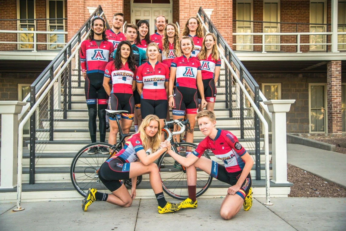 Cycling team standing on Old Main steps, two cyclists in front gripping arms together