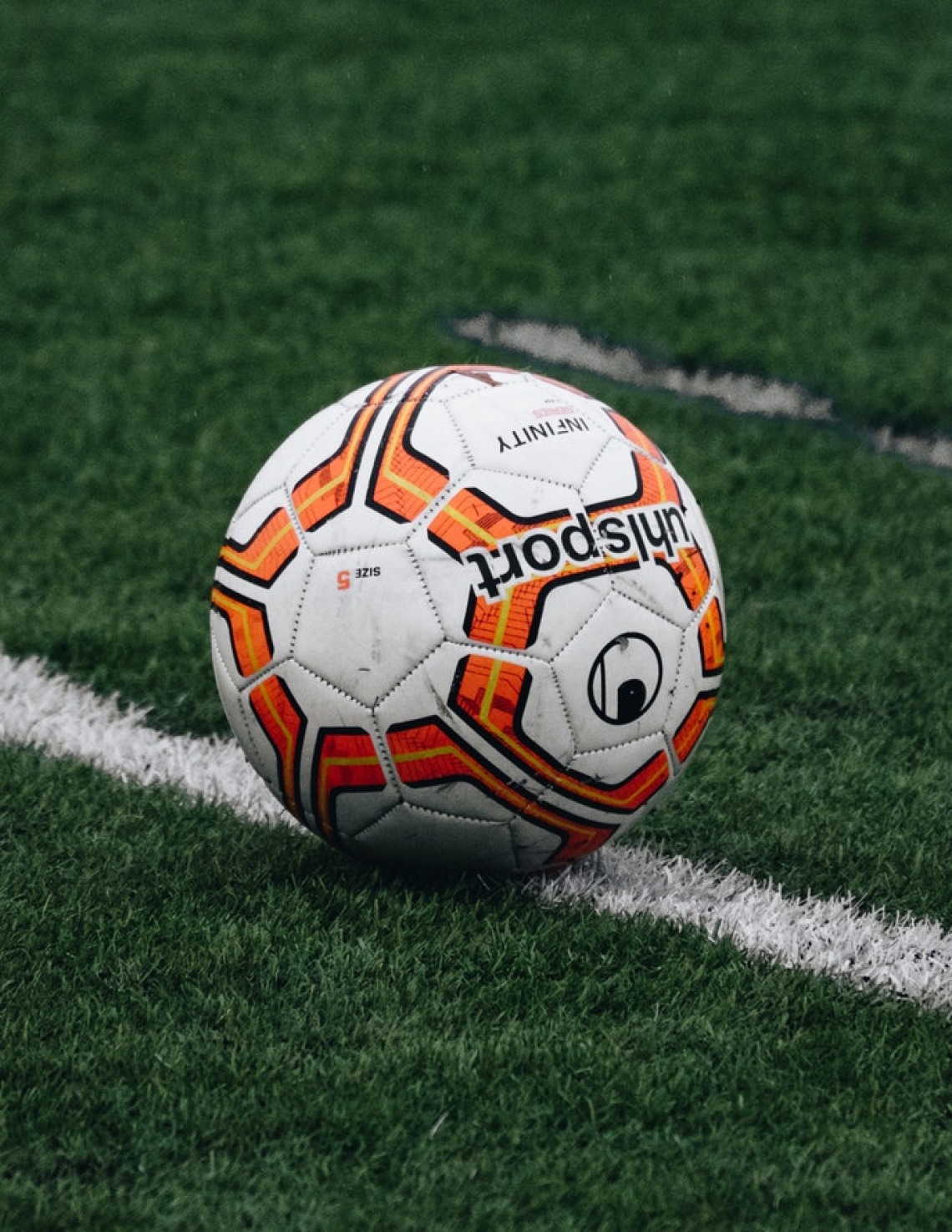 White and orange Uhlsport soccer ball sitting on field next to sideline