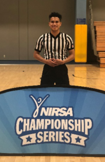 Aguilar wearing sports official uniform, standing behind sign that reads "NIRSA Championship Series"